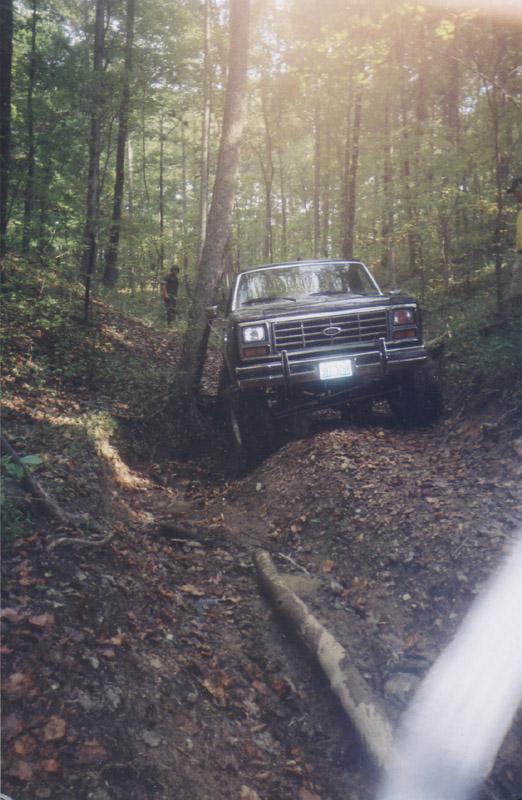 Lifted dark Ford Bronco with brush guard descending a rutted wooded trail, seen from the front