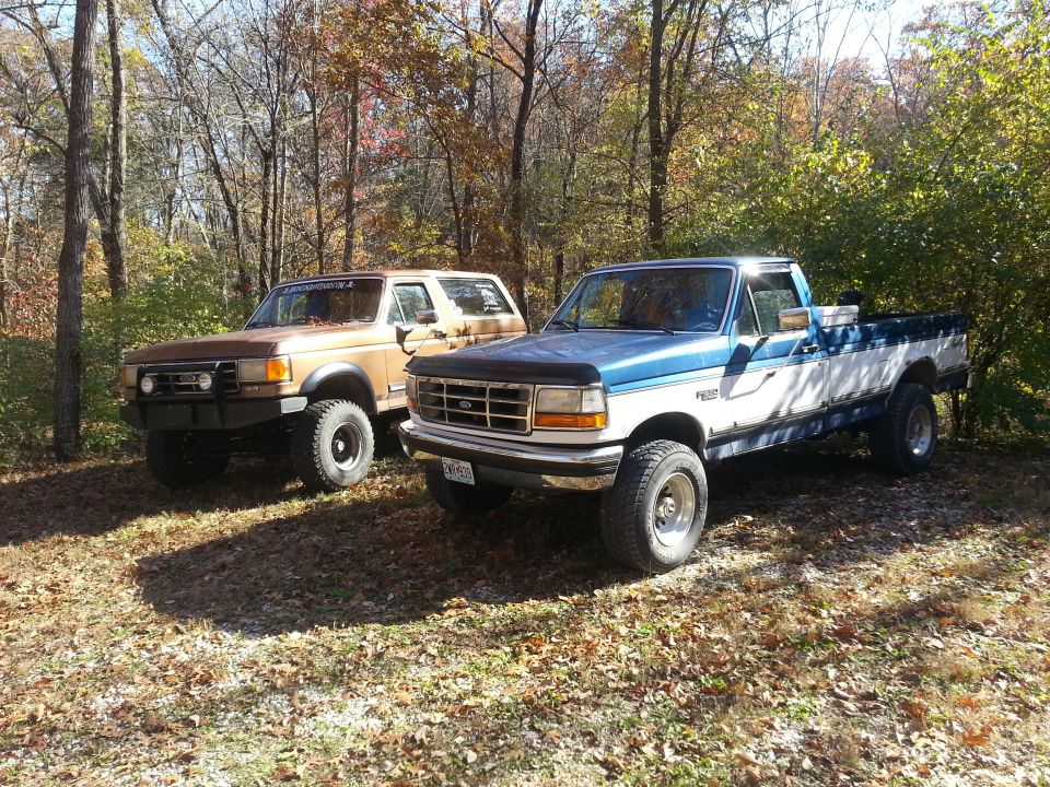 Two pickup trucks, a blue and white Ford and a brown Ford Bronco, parked outdoors in autumn. The blue truck is in the foreground