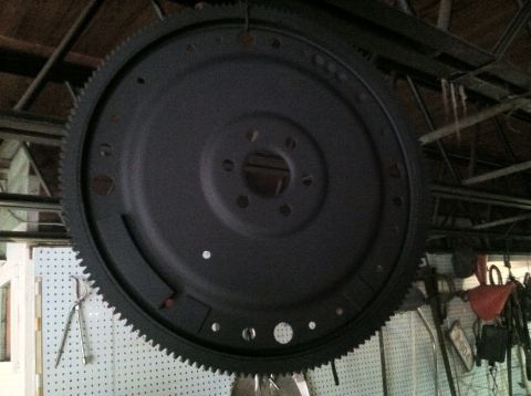 1978 Ford Bronco - A black flywheel with a toothed outer edge is suspended from the ceiling in a workshop.