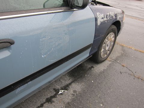2001 Ford Crown Victoria - The side of a blue 2001 Ford Crown Victoria shows significant damage and peeling paint on the front fender.