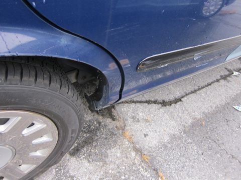 2001 Ford Crown Victoria - A close-up view of a blue sedan's side, revealing scratches and damage to the door and trim, with another vehicle reflected in the paint.