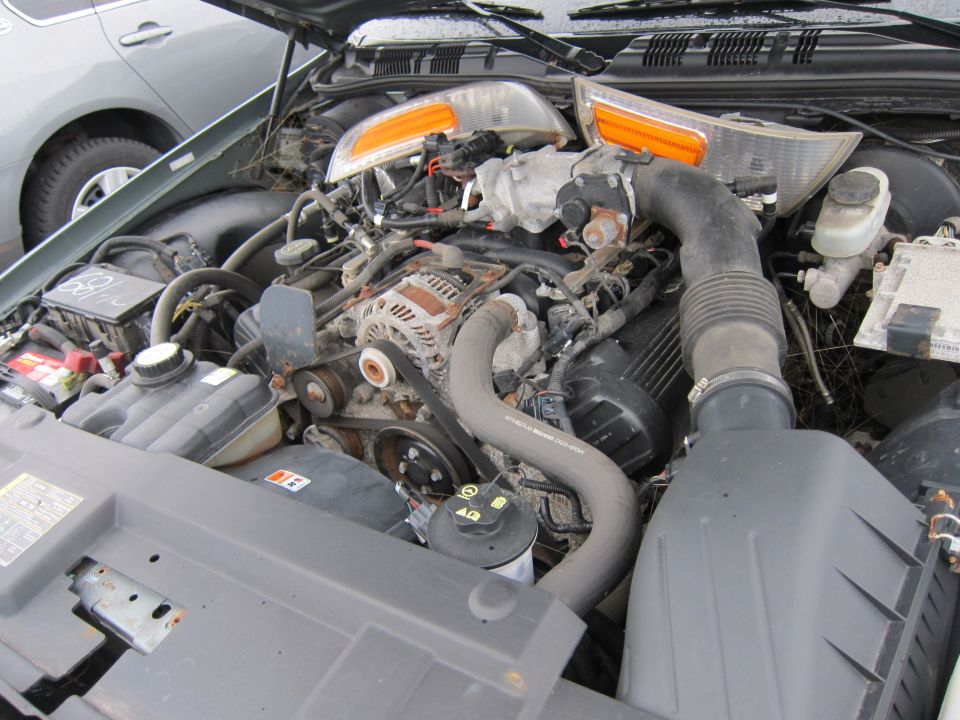Engine bay with exposed components and a grey car in the background