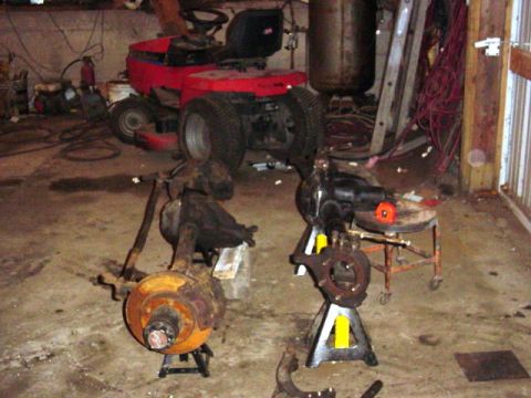 1992 Ford Bronco - Two rusty front axle assemblies are displayed on jack stands in a cluttered garage setting.