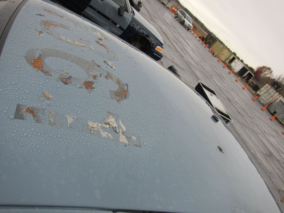 Light blue car hood with peeling decals and water droplets, showing part of a dark car in the background