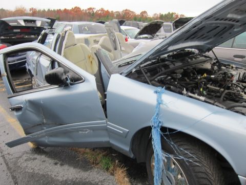 2001 Ford Crown Victoria - A damaged light blue sedan sits in a junkyard with its doors and hood open, showing extensive damage.