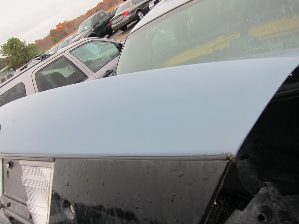 light blue car trunk, black lower body, and a white taillight assembly. Other cars are parked in the background