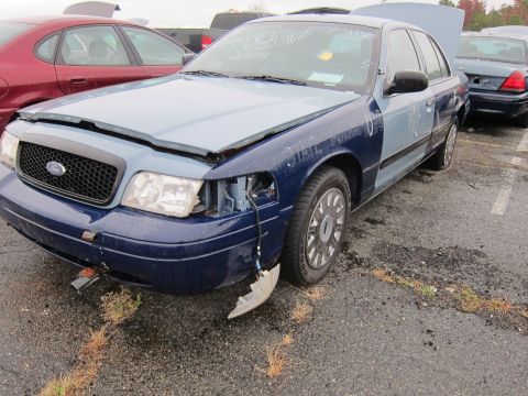 2001 Ford Crown Victoria - A damaged 2001 Ford Crown Victoria sits in a lot with other vehicles, its hood and one headlight askew.