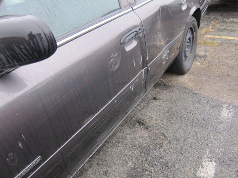 2001 Ford Crown Victoria - A dark sedan with visible damage to its side, including dents and scratches on the rear door and fender.
