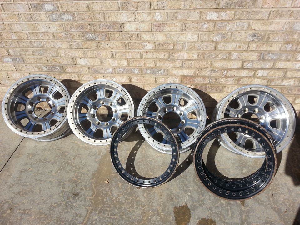 Four aluminum wheels and two beadlock rings against a brick wall