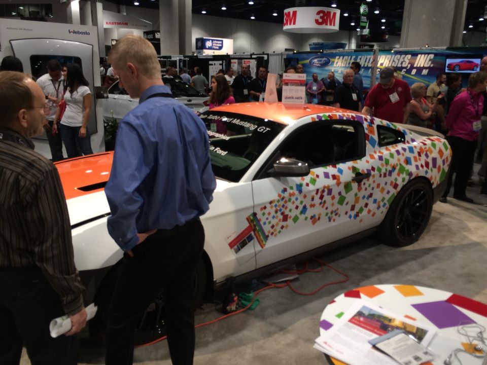 White 2013 Ford Mustang GT with orange hood and colorful square vinyl wrap, parked indoors at a show