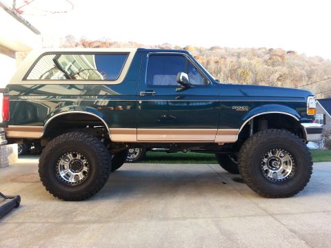 1995 Ford Bronco - The side profile of a dark green 1995 Ford Bronco is shown, lifted with large off-road tires and custom silver rims. The vehicle has a tan…