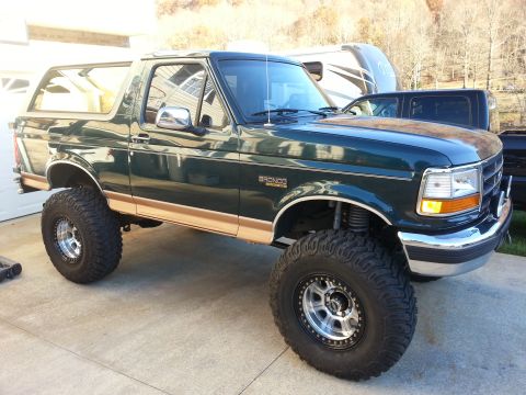 1995 Ford Bronco - A lifted, dark green 1995 Ford Bronco with tan lower trim and chrome accents is parked on a concrete driveway in front of a garage. The veh…