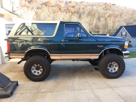 1995 Ford Bronco - A lifted, dark green 1995 Ford Bronco with a tan top is parked on a concrete driveway. The vehicle has large off-road tires and custom rims.