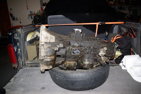 1995 Ford Bronco - A dirty automatic transmission is resting on a tire in the bed of a dark blue SUV. The transmission is secured with orange straps, and othe…