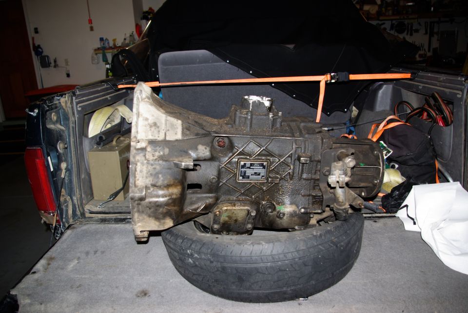 Dirty automatic transmission on a tire in the bed of a dark blue SUV, secured with orange straps.