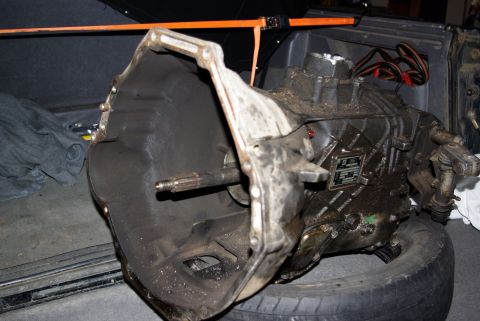 1995 Ford Bronco - A dirty, greasy Ford transmission is resting on a tire inside what appears to be the cargo area of a vehicle. An orange strap is visible ov…