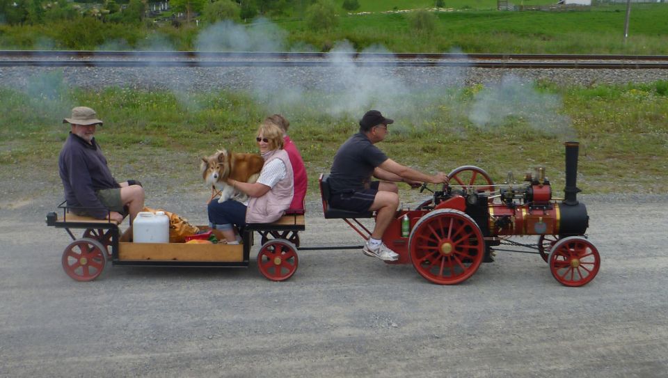 Miniature red and black steam engine pulling a wagon with people and a dog.