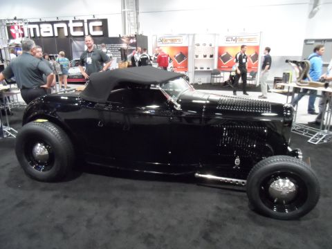 1971 Ford Ranchero - A shiny black convertible hot rod is displayed on a black carpeted floor at an indoor event with booths and people in the background.
