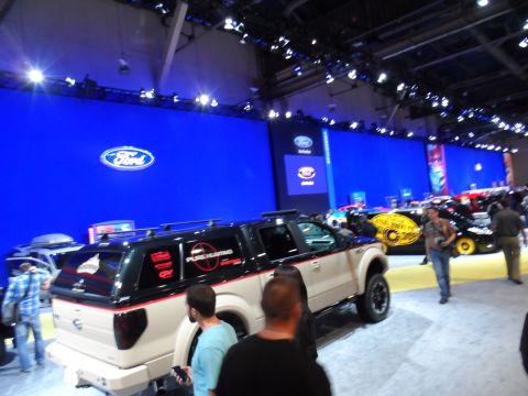 1971 Ford Ranchero - A lifted, two-tone beige and black Ford pickup truck with red accents is on display at an auto show. The truck is parked in front of a larg…