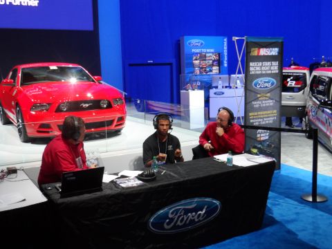 1971 Ford Ranchero - A red Ford Mustang is displayed on a platform with two men wearing headphones sitting at a table in front of it. A NASCAR banner is visible…