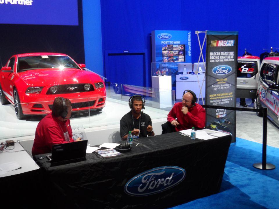 Red Ford Mustang on display with two men at a table and a NASCAR banner.