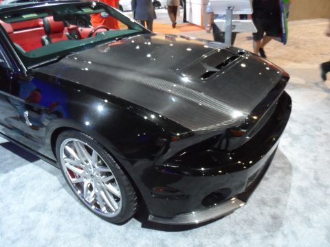 1971 Ford Ranchero - The front passenger side of a black convertible sports car with a carbon fiber hood is shown at an indoor event. The car has red interior s…