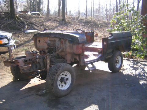 1978 Ford Bronco - A stripped 4x4 truck body shell sitting outdoors on a gravel driveway in a wooded lot, viewed from the front three-quarter with the front c…
