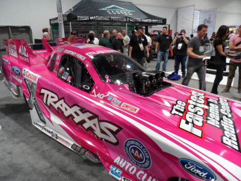 1971 Ford Ranchero - A bright pink drag racing car with "TRAXXAS" and "The Fastest Name In Radio Control" text is displayed indoors at an event. Several people…
