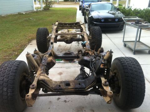 1979 Ford Bronco - The chassis and frame of a vehicle are visible, with a black Ford Mustang and other vehicles in the background.