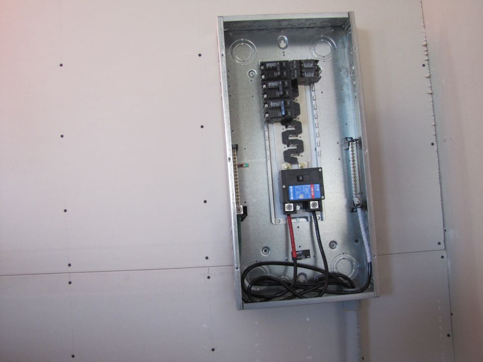 Open electrical panel box with circuit breakers and wiring on a drywall wall.