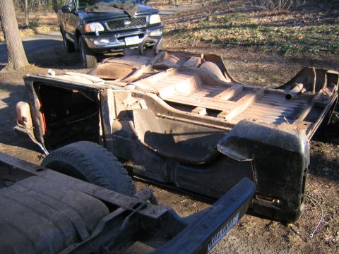 1978 Ford Bronco - The chassis of a vehicle sits outdoors, with a dark pickup truck parked behind it.