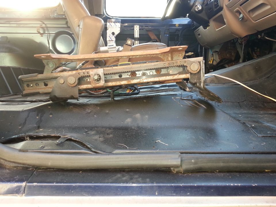 Interior of a blue vehicle with the driver's seat removed, showing exposed floor and seat rails