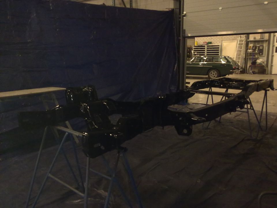 Black painted vehicle frame on stands in a garage, with a green station wagon visible in the background