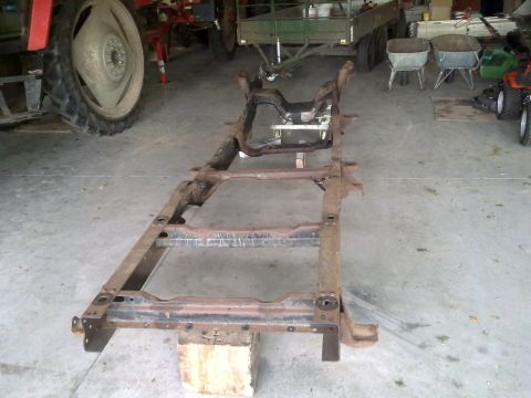 1984 Ford Bronco - A vehicle frame sits on wooden blocks, undergoing what appears to be a chassis rebuild, with farm equipment visible in the background.
