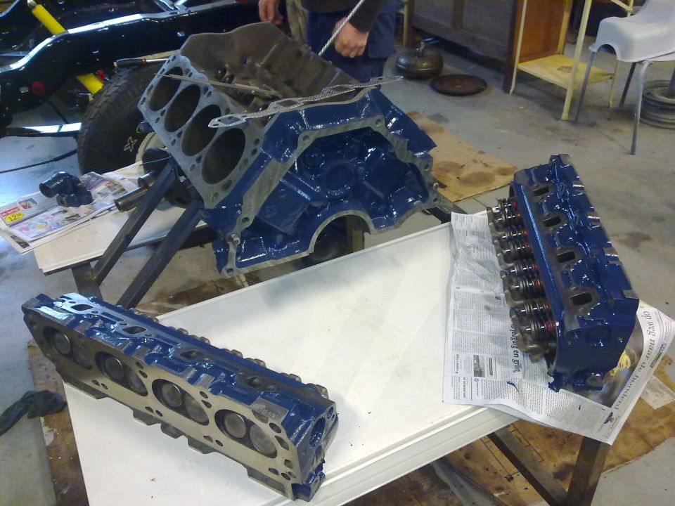 Engine block and two cylinder heads, all painted blue, sit on a table in a garage. A vehicle frame is visible in the background