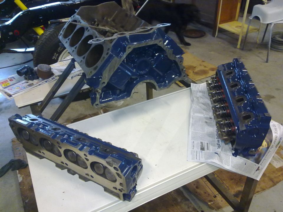 Blue engine block and two cylinder heads on a table, with a partial vehicle in the background