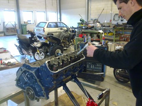 1984 Ford Bronco - Mechanic works on an engine block in a garage filled with various vehicles and equipment.