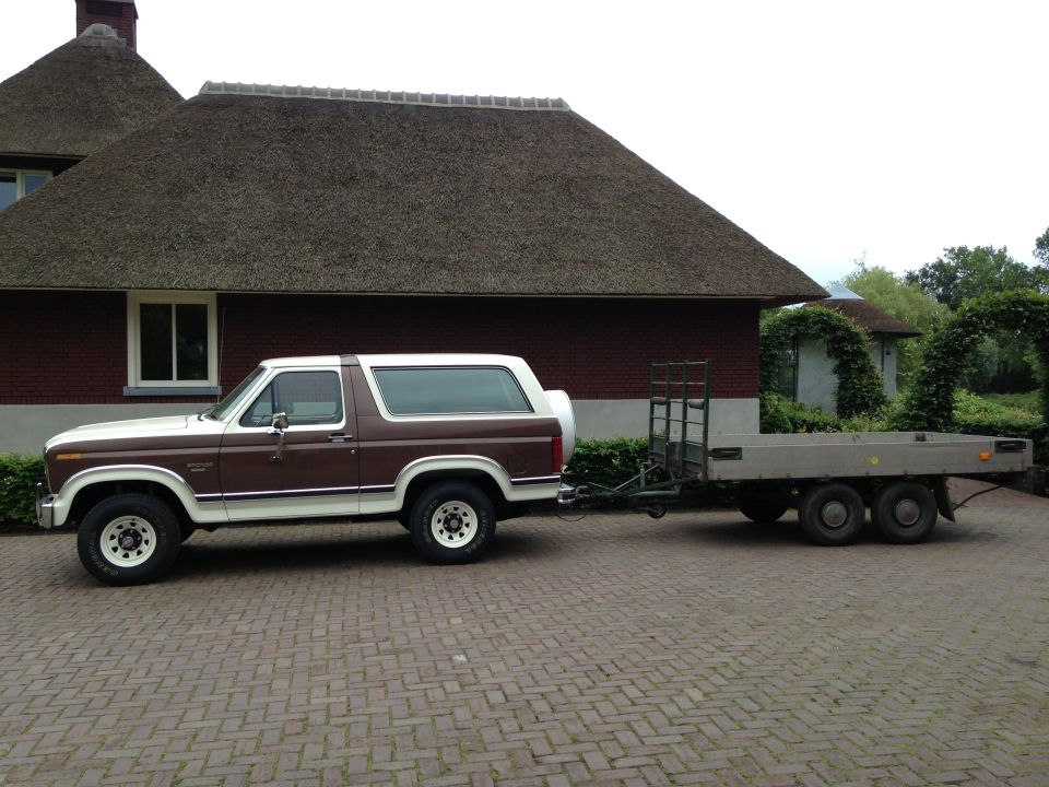 Brown and white Ford Bronco with a spare tire on the back, towing a flatbed trailer