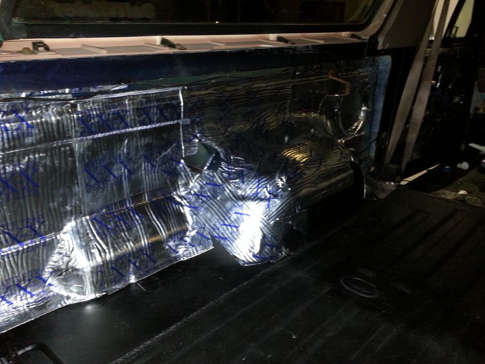 Interior of a vehicle with silver and blue sound dampening material covering the rear side panel and black carpeted floor