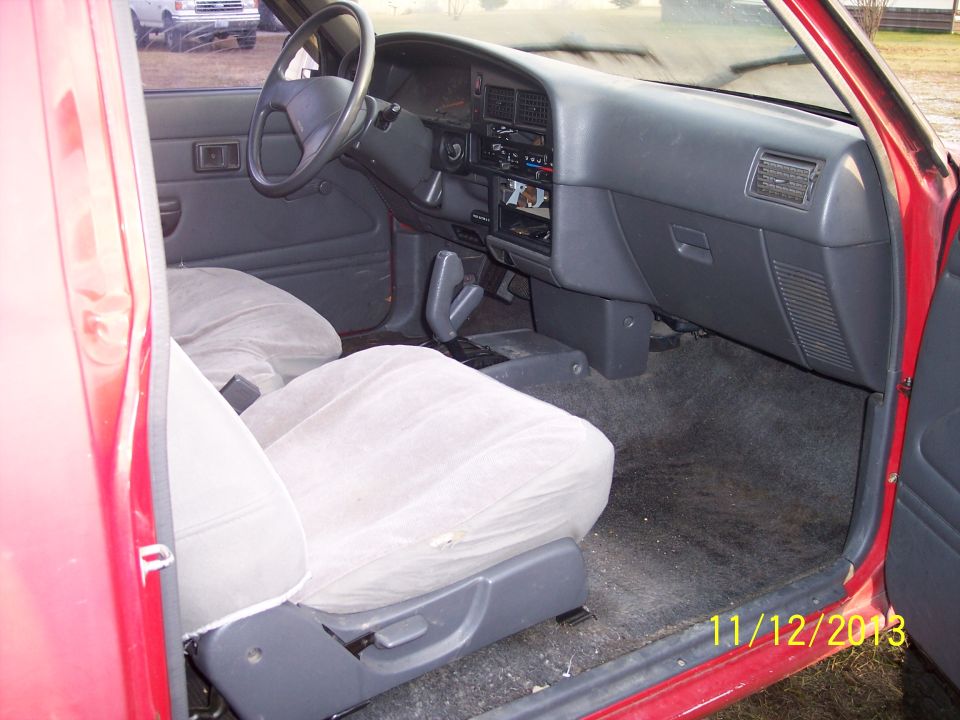 Interior of a red truck, showing the dashboard, steering wheel, and seats. Another white truck is visible outside