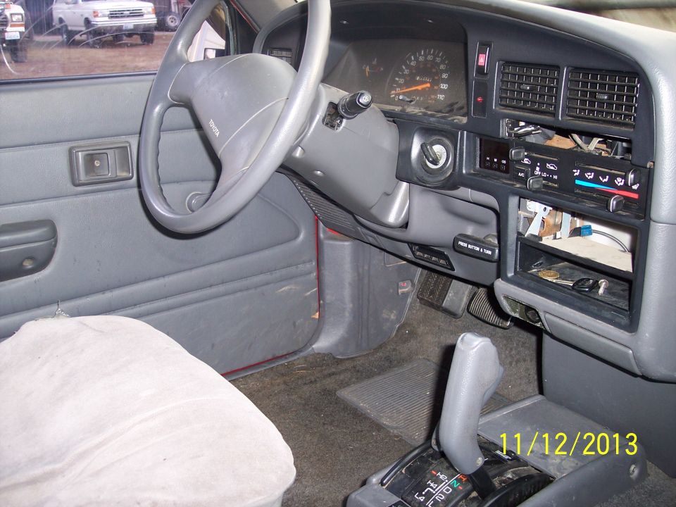 Interior of a red Toyota pickup truck, showing the dashboard, steering wheel, and gear shifter. A white pickup truck is visible outside the driver's side window