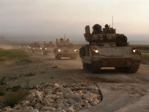 1982 Ford Bronco - A convoy of armored vehicles moves along a dusty road as the sun sets.