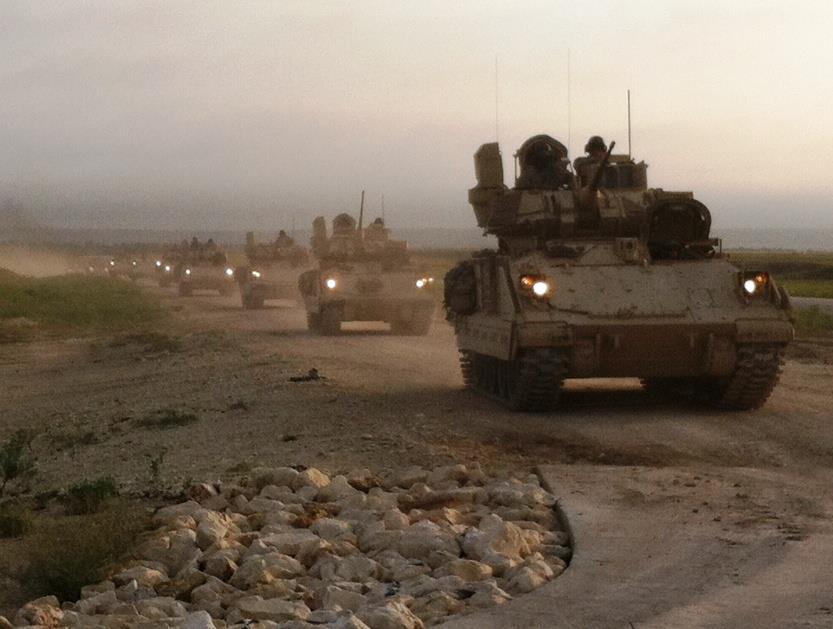 Column of armored military vehicles driving on a dirt road at dusk