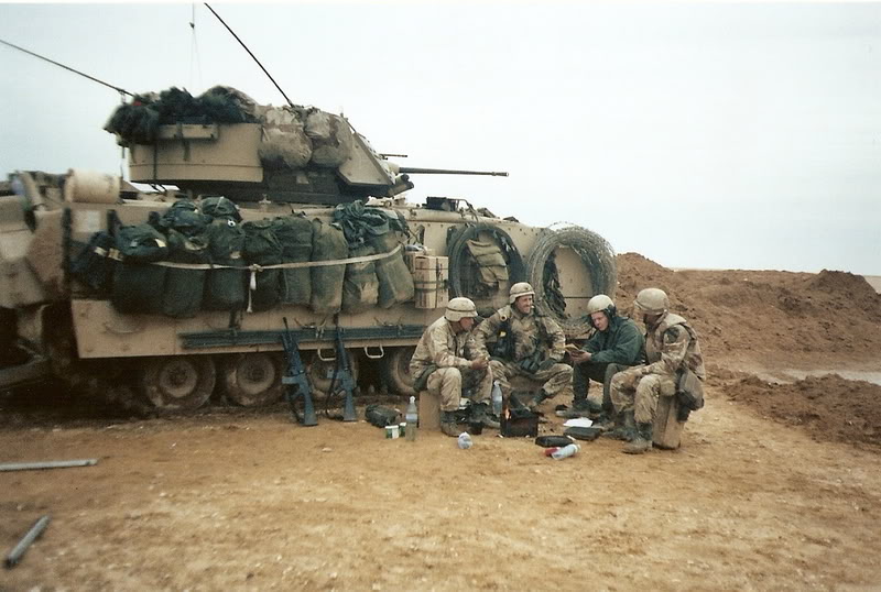 Side view of a tan military vehicle with soldiers sitting in front of it