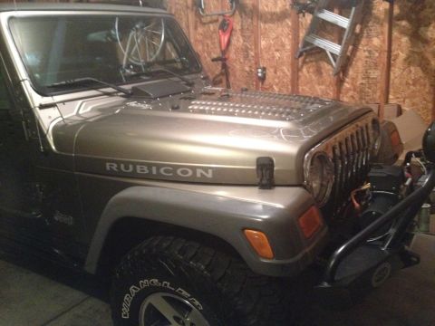 2005 Jeep Wrangler - Tan Jeep Wrangler Rubicon with louvered hood, seven-slot grille and Wrangler tires parked inside an OSB-walled garage.