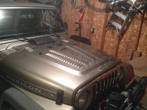 2005 Jeep Wrangler - The hood of a gold Jeep Wrangler, identified by the 'Rubicon' decal, is shown with a vented insert and chrome grille details.