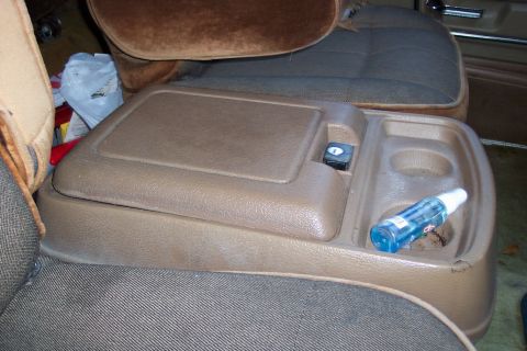 1986 Ford Bronco - The interior of a vehicle shows a brown center console with a storage compartment and cup holders, alongside fabric seats.