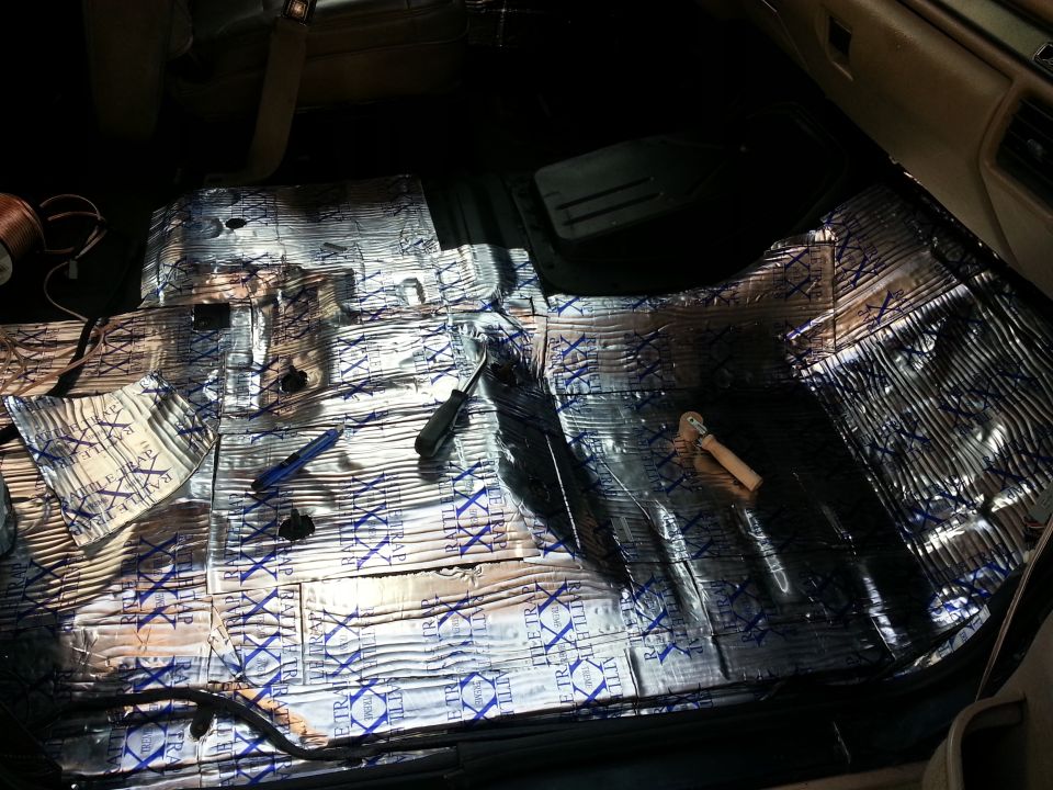 Car floor covered in silver sound deadening material with blue text. Screwdriver and pliers visible