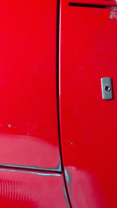 1964 Ford Fairlane - Detail of a red car's body panel seam, showing a small metal tab on the right side. The exact make and model are not identifiable from this…