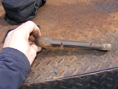 Rusty metal tool with a pointed end and a notch, held by a hand on a rusty surface.